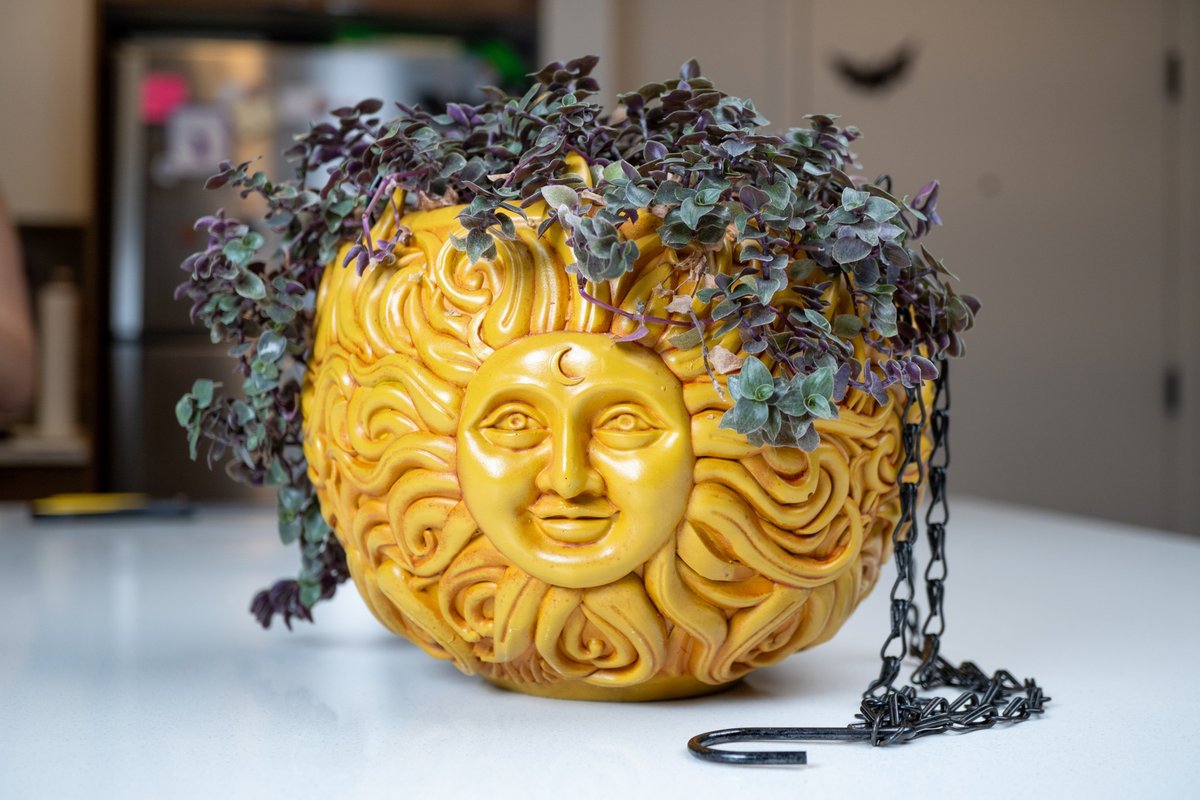 Unedited camera photo of the mustard-yellow resin sun face planter on a counter, its chain beside it